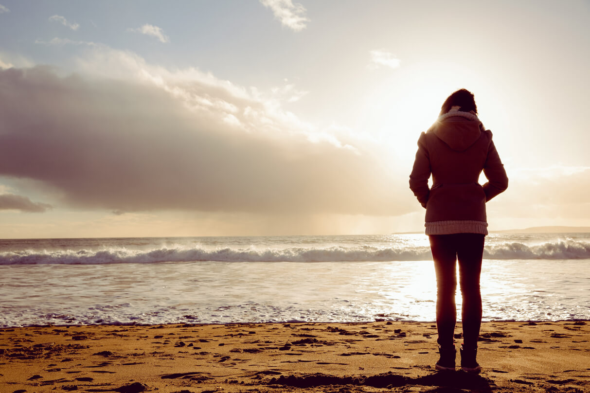 woman stands on beach in coat watching waves